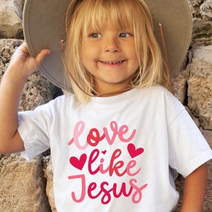 Peut inclure: Un t-shirt blanc avec le texte "love like Jesus" en lettres roses et rouges avec des c&oelig;urs roses.