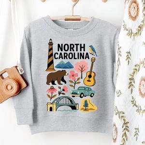 May include: A light gray sweatshirt featuring a colorful graphic of North Carolina landmarks and symbols. The design includes a lighthouse, bear, mountains, flowers, a guitar, a car, a bridge, and a bird. The words "NORTH CAROLINA" are printed in black.