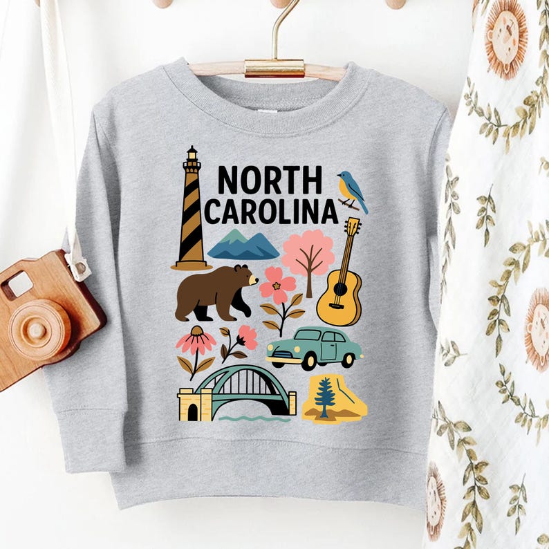 May include: A light gray sweatshirt featuring a colorful graphic of North Carolina landmarks and symbols. The design includes a lighthouse, bear, mountains, flowers, a guitar, a car, a bridge, and a bird. The words "NORTH CAROLINA" are printed in black.