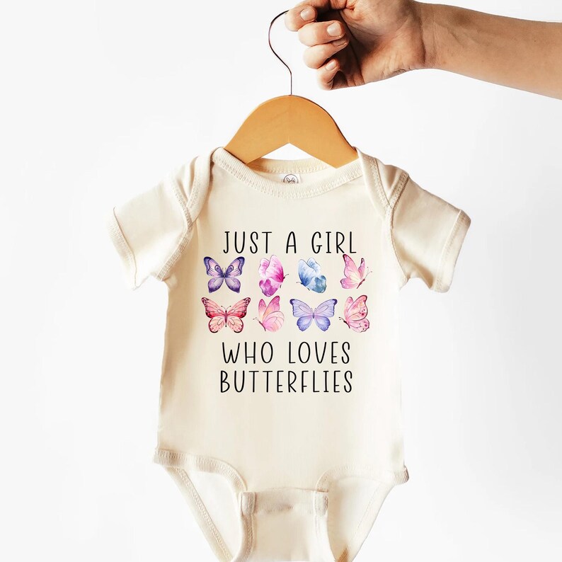 May include: Cream-colored baby bodysuit with the text "JUST A GIRL WHO LOVES BUTTERFLIES." The bodysuit features a collection of watercolor butterflies in shades of purple, pink, and blue. The garment is hanging on a wooden hanger.