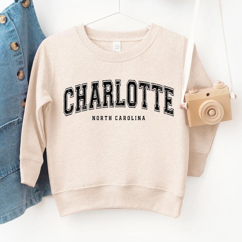 May include: A beige sweatshirt with the word "CHARLOTTE" in black, outlined letters, and "NORTH CAROLINA" below it. The sweatshirt is hanging on a white hanger. A toy camera hangs to the right.
