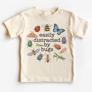 Peut inclure: T-shirt crème avec un assortiment coloré d'insectes illustrés, dont une coccinelle, une abeille et un papillon. Le texte "easily distracted by bugs" est centré sur le t-shirt. Le t-shirt est en matière douce.
