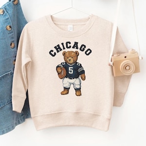 May include: A beige sweatshirt with the word "CHICAGO" arched across the top. A cartoon bear wearing a navy blue football jersey with the number 5 and holding a football is in the center. The sweatshirt is hanging on a white hanger.