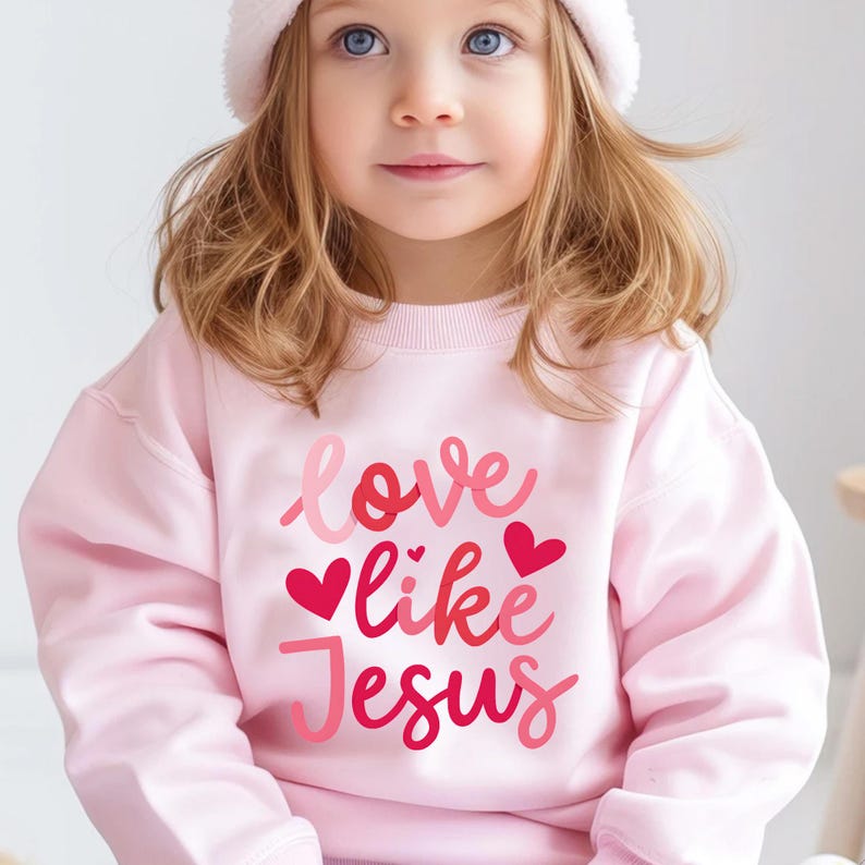 Peut inclure: Un sweat-shirt rose avec le texte "love like Jesus" en lettres roses, rouges et blanches. Il y a trois petits c&oelig;urs roses autour du texte.