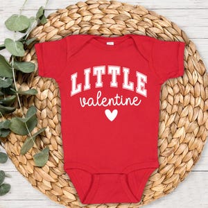 May include: A red baby bodysuit with the words "LITTLE valentine" in white, with a white heart below the text. The bodysuit is short-sleeved and made of a soft material. The bodysuit is displayed on a woven, circular mat with green leaves.