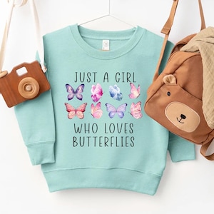 May include: A light blue sweatshirt with the text "JUST A GIRL WHO LOVES BUTTERFLIES" and colorful butterfly graphics. A wooden toy camera and a brown bear backpack hang on either side of the sweatshirt.