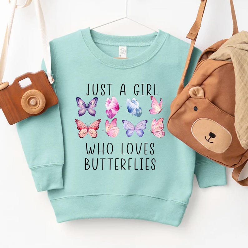 May include: A light blue sweatshirt with the text "JUST A GIRL WHO LOVES BUTTERFLIES" and colorful butterfly graphics. A wooden toy camera and a brown bear backpack hang on either side of the sweatshirt.