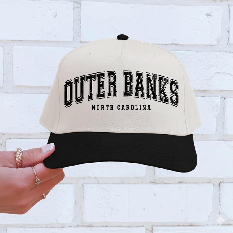 May include: A cream-colored baseball cap with a black brim and a black button on top. The cap features the text "OUTER BANKS NORTH CAROLINA" in a bold, black, collegiate-style font.