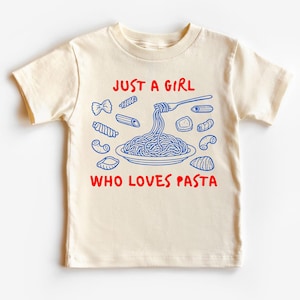 Op de afbeelding: Cr&egrave;mekleurig peuter-T-shirt met een blauwe en rode afbeelding. De afbeelding toont verschillende pastavormen rond een bord spaghetti dat door een vork wordt opgetild. De tekst luidt "JUST A GIRL WHO LOVES PASTA."