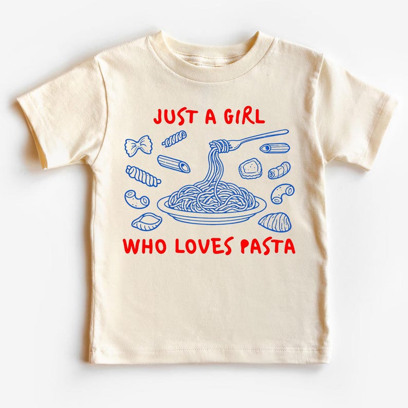 Op de afbeelding: Cr&egrave;mekleurig peuter-T-shirt met een blauwe en rode afbeelding. De afbeelding toont verschillende pastavormen rond een bord spaghetti dat door een vork wordt opgetild. De tekst luidt "JUST A GIRL WHO LOVES PASTA."