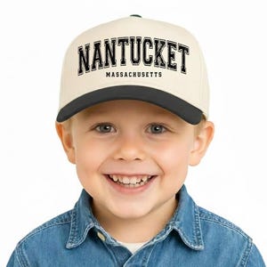 Può includere: Un cappellino da baseball color crema con visiera nera, con la scritta "NANTUCKET" in grassetto nero. Sotto, "MASSACHUSETTS" in lettere nere più piccole. Il cappellino è indossato da un bambino sorridente con una camicia di jeans.