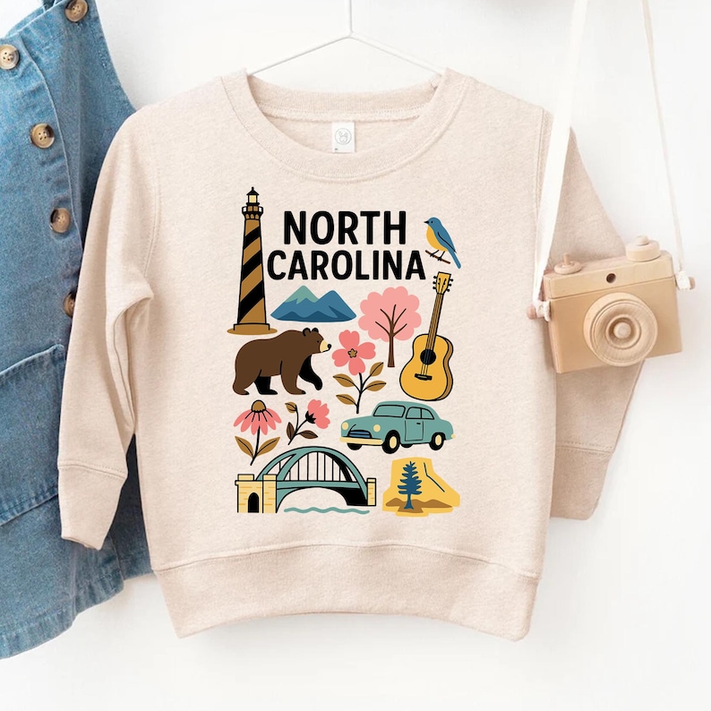 May include: A beige sweatshirt featuring a colorful graphic of North Carolina landmarks. The design includes a lighthouse, mountains, a bear, flowers, a guitar, a car, and the text "NORTH CAROLINA".
