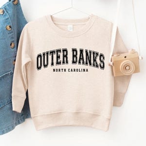 May include: A beige sweatshirt with the text "OUTER BANKS NORTH CAROLINA" in black block letters. The sweatshirt is hanging on a white hanger. A small toy camera hangs from the hanger.