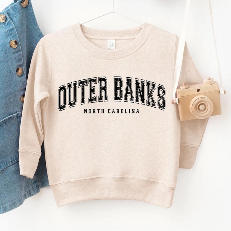 May include: A beige sweatshirt with the text "OUTER BANKS NORTH CAROLINA" in black block letters. The sweatshirt is hanging on a white hanger. A small toy camera hangs from the hanger.