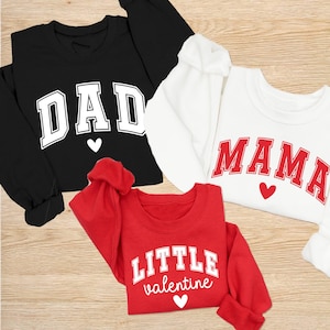 May include: Three sweatshirts: black with "DAD", white with "MAMA", and red with "LITTLE valentine". Each has a heart graphic. The text is in a collegiate style font. The sweatshirts are long-sleeved and made of a soft material.
