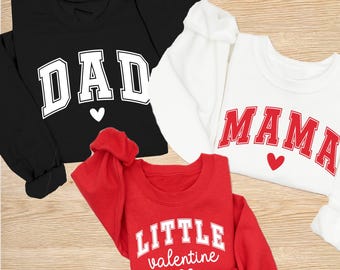 Peut inclure: Trois sweatshirts : noir avec "DAD", blanc avec "MAMA" et rouge avec "LITTLE valentine". Chacun a un cœur. Le texte est dans une police de style universitaire. Les sweatshirts sont à manches longues et en matière douce.