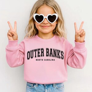 May include: A light pink sweatshirt with the text "OUTER BANKS NORTH CAROLINA" in black. The sweatshirt is worn by a person wearing white heart-shaped sunglasses and making a peace sign with both hands.