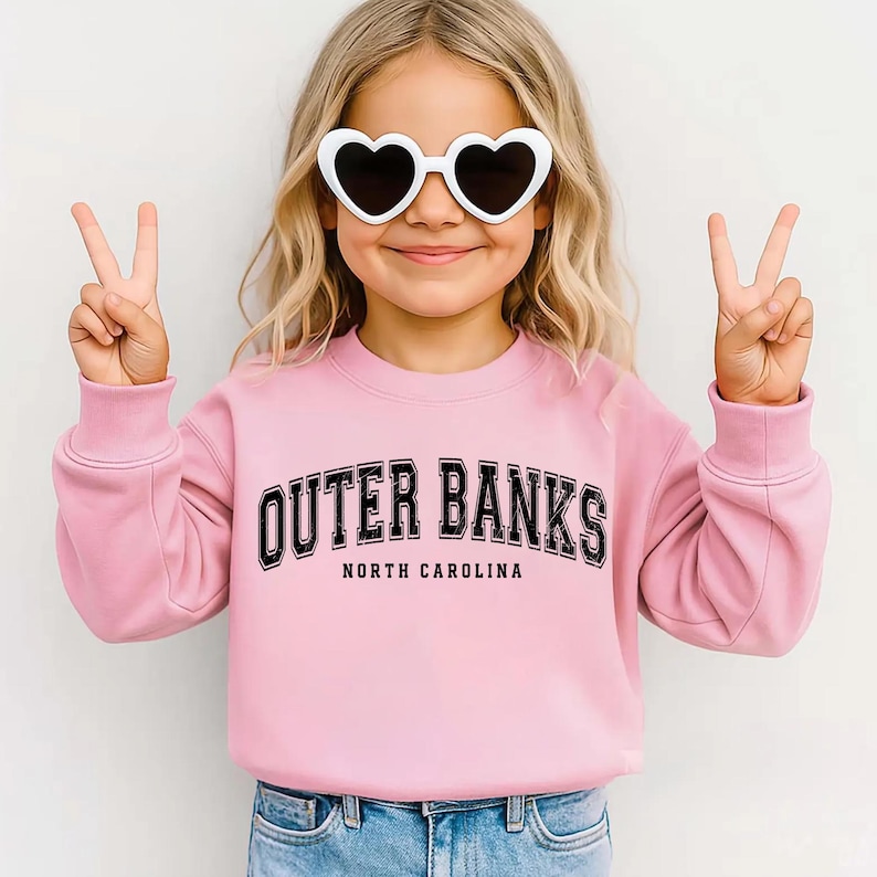 May include: A light pink sweatshirt with the text "OUTER BANKS NORTH CAROLINA" in black. The sweatshirt is worn by a person wearing white heart-shaped sunglasses and making a peace sign with both hands.