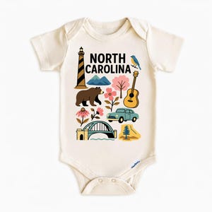 May include: Cream-colored baby bodysuit with a colorful graphic of North Carolina landmarks, including a lighthouse, mountains, bear, guitar, car, and bridge. The words "North Carolina" are printed in black.