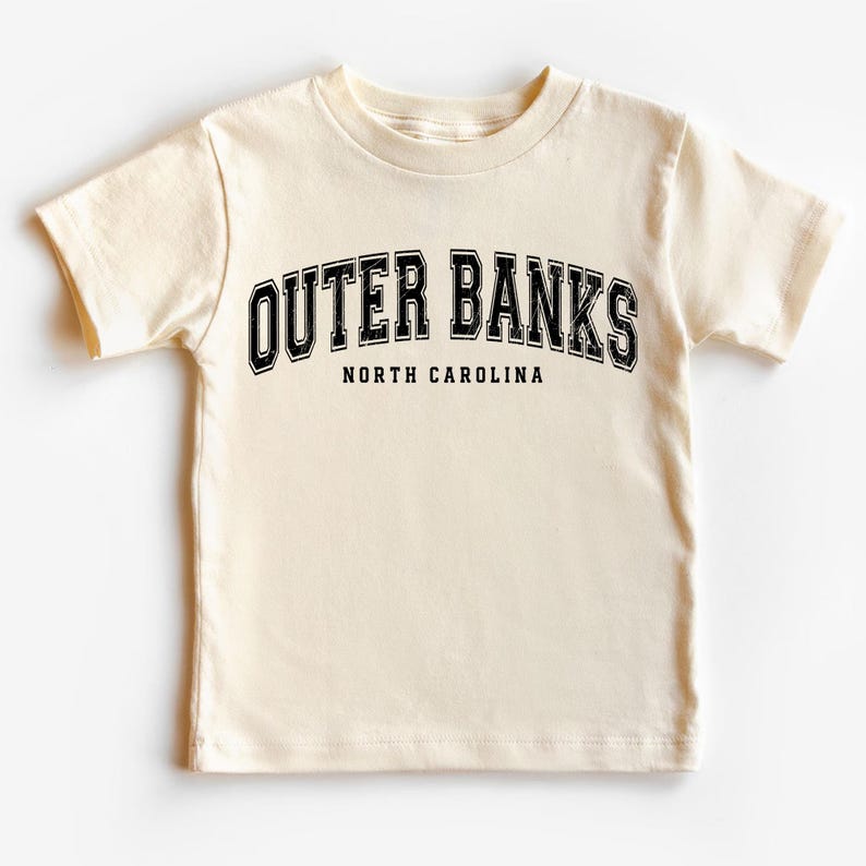 May include: Cream-colored t-shirt featuring the text "OUTER BANKS" in a black, arched font above "NORTH CAROLINA". The shirt has short sleeves and a crew neck. A casual, everyday wear tee.