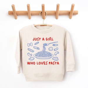 Op de afbeelding: Een beige sweatshirt met de tekst "JUST A GIRL WHO LOVES PASTA" in rood en blauw. Het ontwerp toont verschillende pastavormen en een vork die spaghetti oprolt. De sweatshirt hangt aan een houten kledingrek.