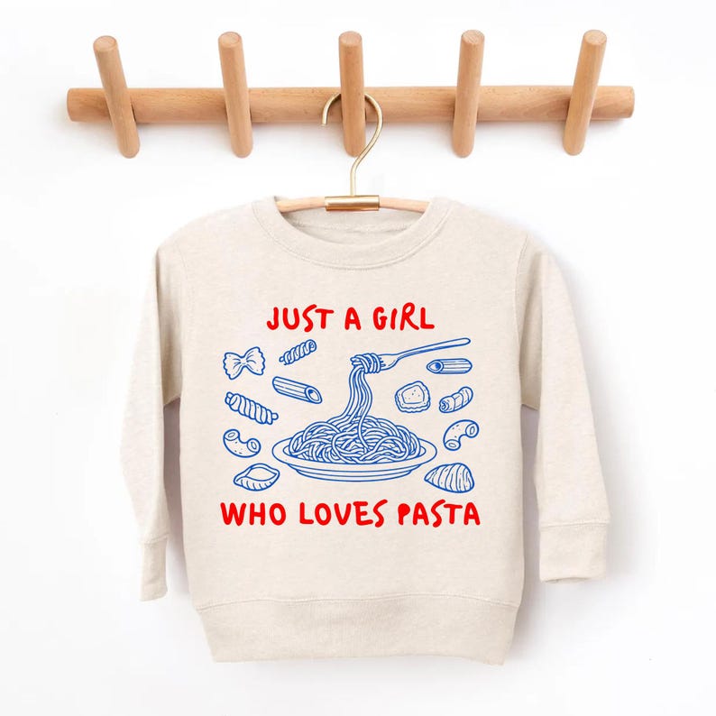 Op de afbeelding: Een beige sweatshirt met de tekst "JUST A GIRL WHO LOVES PASTA" in rood en blauw. Het ontwerp toont verschillende pastavormen en een vork die spaghetti oprolt. De sweatshirt hangt aan een houten kledingrek.