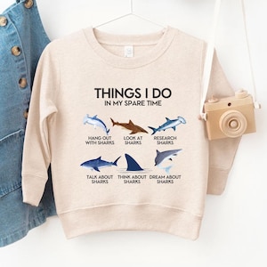 Puede incluir: Sudadera beige con el texto "THINGS I DO IN MY SPARE TIME" e ilustraciones de tiburones. El diseño incluye frases como "Hang out with sharks" y "Dream about sharks."