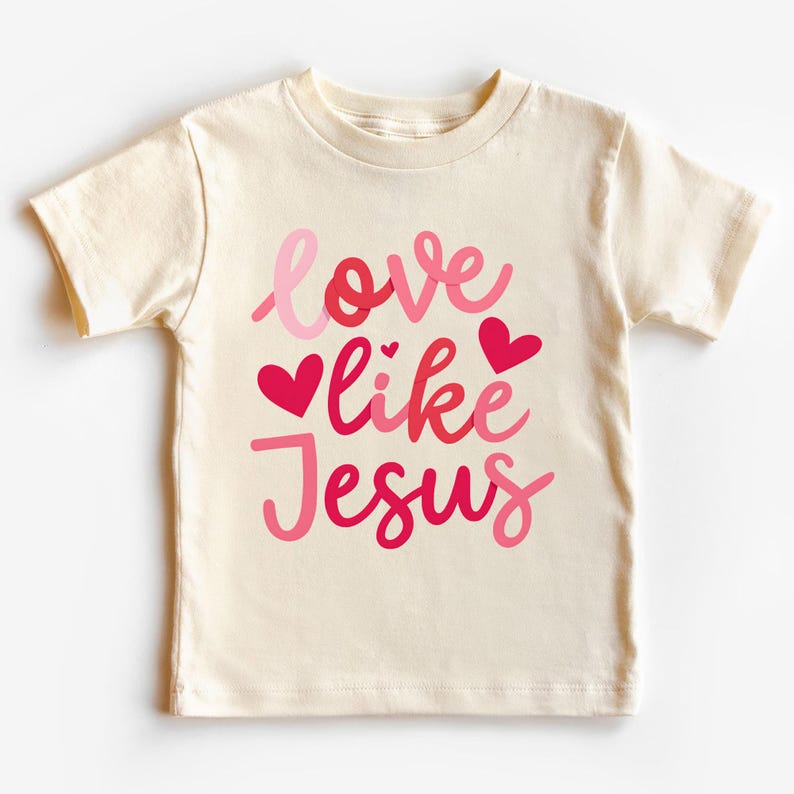 Peut inclure: Un t-shirt cr&egrave;me avec le texte "love like Jesus" en lettres roses et rouges avec des c&oelig;urs roses.