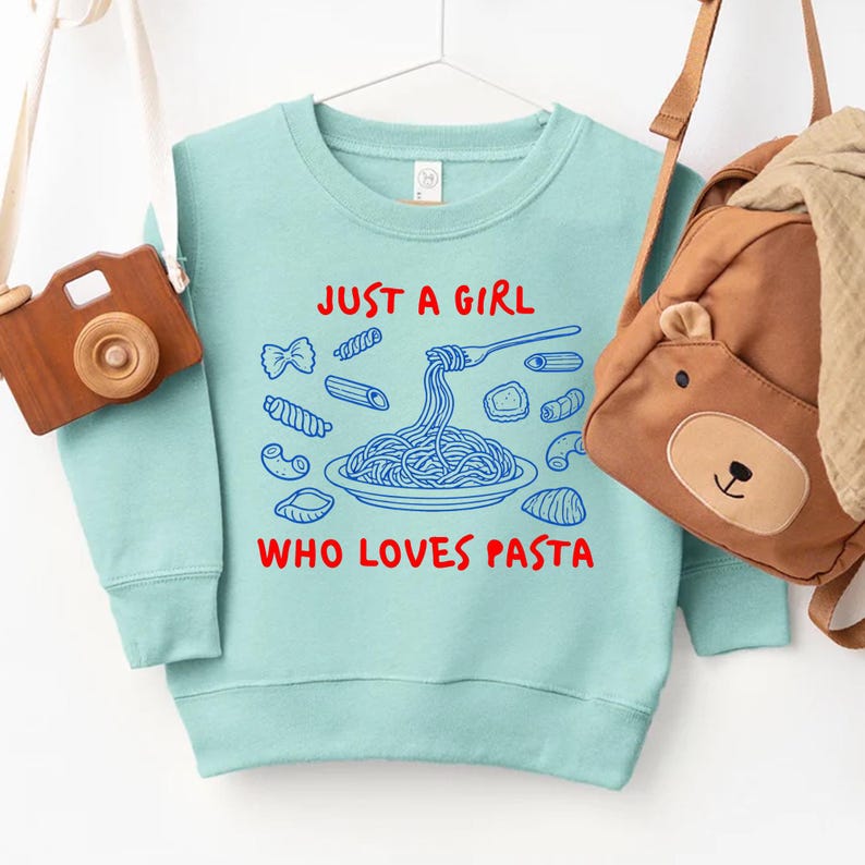 Op de afbeelding: Een lichtblauw sweatshirt met de tekst "JUST A GIRL WHO LOVES PASTA" in rood en blauw. De afbeelding toont een bord spaghetti en verschillende pastavormen. Een bruine berenrugzak en een houten camera zijn zichtbaar.