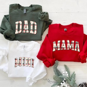 May include: Four sweatshirts in the image: a green one with "DAD", a red one with "MAMA", and a white one with "MINI". The text is in a plaid pattern. The sweatshirts are on a light wood surface.