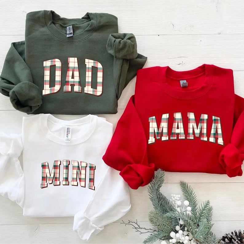 May include: Four sweatshirts in the image: a green one with "DAD", a red one with "MAMA", and a white one with "MINI". The text is in a plaid pattern. The sweatshirts are on a light wood surface.