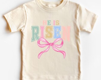 May include: A light beige t-shirt with a pastel rainbow colored graphic that says "He is Risen" with a pink bow underneath.