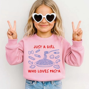 Op de afbeelding: Een roze sweatshirt met de tekst "JUST A GIRL WHO LOVES PASTA" in rood en blauw. De afbeelding toont verschillende pastavormen en een bord spaghetti. De persoon op de foto draagt een hartvormige zonnebril en maakt een vredesteken.