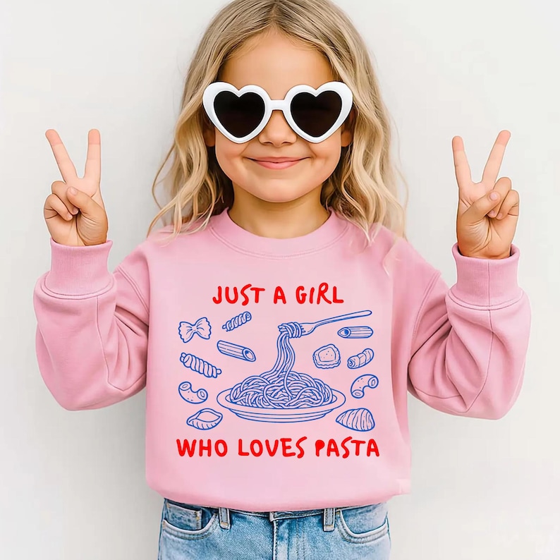 Op de afbeelding: Een roze sweatshirt met de tekst "JUST A GIRL WHO LOVES PASTA" in rood en blauw. De afbeelding toont verschillende pastavormen en een bord spaghetti. De persoon op de foto draagt een hartvormige zonnebril en maakt een vredesteken.