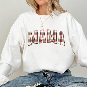 May include: White sweatshirt with the word "MAMA" in a plaid pattern across the chest. The plaid design incorporates red, green, and brown colors. The sweatshirt is a crew neck style.