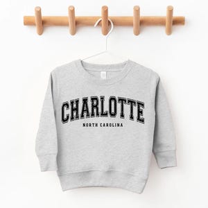 May include: A light gray sweatshirt with the word "CHARLOTTE" in black, arched lettering. Below the word "CHARLOTTE" is the text "NORTH CAROLINA" in a smaller, straight font. The sweatshirt is hanging on a white hanger from a wooden rack.