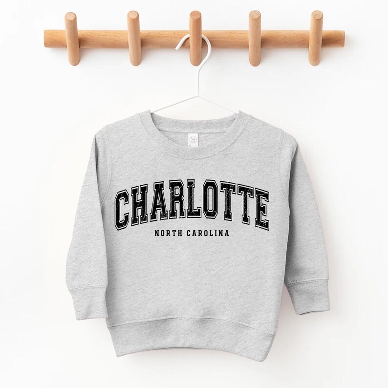 May include: A light gray sweatshirt with the word "CHARLOTTE" in black, arched lettering. Below the word "CHARLOTTE" is the text "NORTH CAROLINA" in a smaller, straight font. The sweatshirt is hanging on a white hanger from a wooden rack.