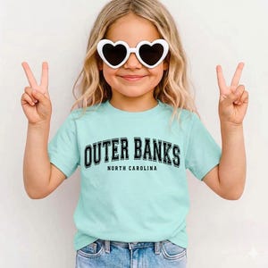 May include: A light blue t-shirt with the text "Outer Banks North Carolina" in black. The person is wearing white heart-shaped sunglasses and making a peace sign with their fingers.