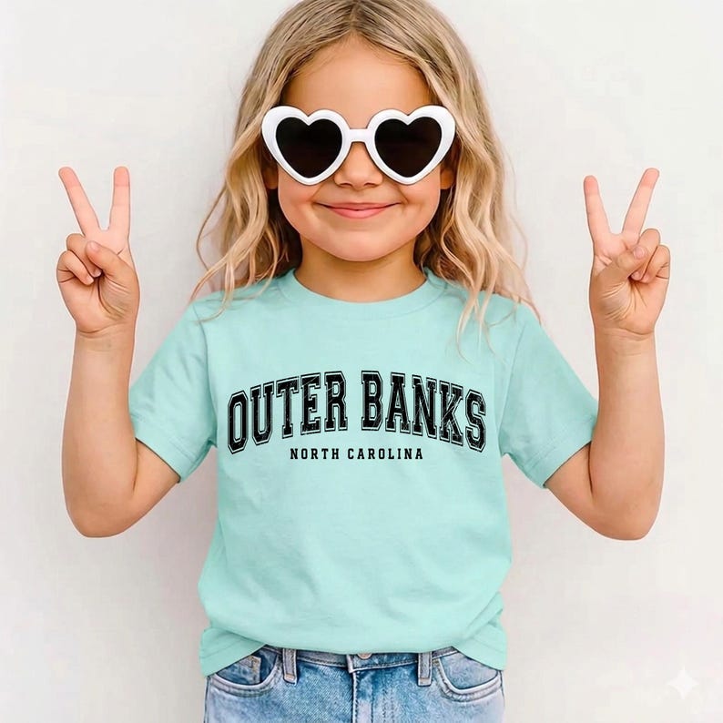 May include: A light blue t-shirt with the text "Outer Banks North Carolina" in black. The person is wearing white heart-shaped sunglasses and making a peace sign with their fingers.