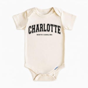 May include: Cream-colored baby onesie with the word "CHARLOTTE" in black, arched text above "NORTH CAROLINA". The onesie has short sleeves and a snap closure at the bottom. The Gerber brand logo is visible.