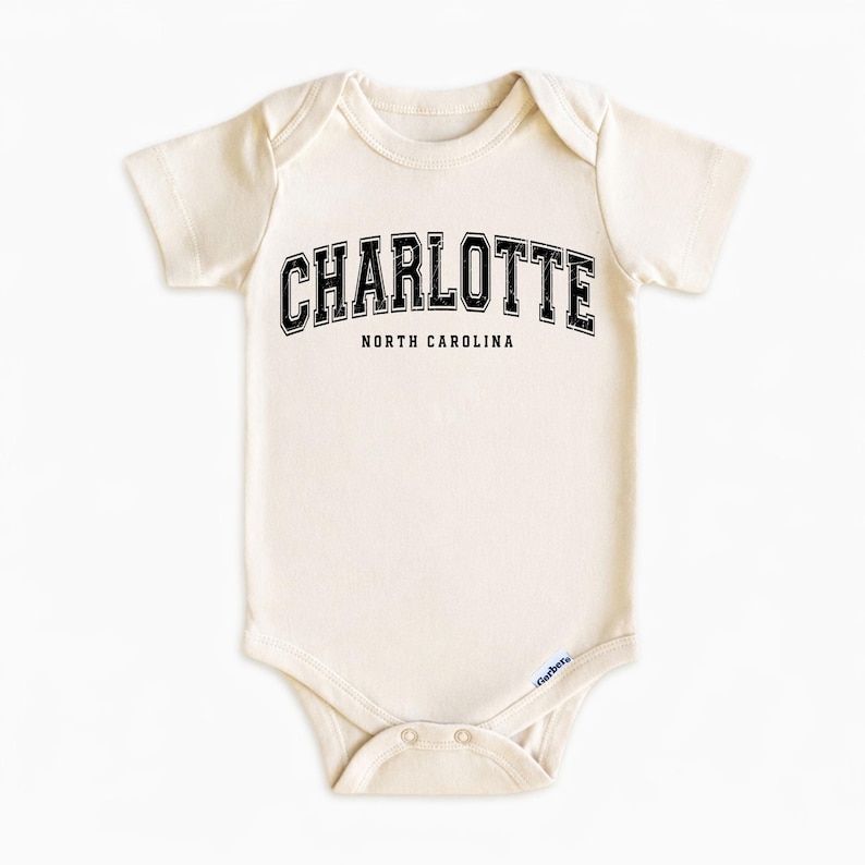 May include: Cream-colored baby onesie with the word "CHARLOTTE" in black, arched text above "NORTH CAROLINA". The onesie has short sleeves and a snap closure at the bottom. The Gerber brand logo is visible.