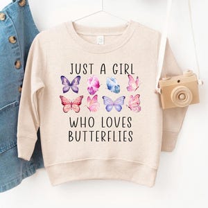 May include: A beige sweatshirt with the text "JUST A GIRL WHO LOVES BUTTERFLIES" in black. The design includes nine colorful butterfly illustrations in shades of purple, pink, and blue. The sweatshirt is hanging on a white hanger.