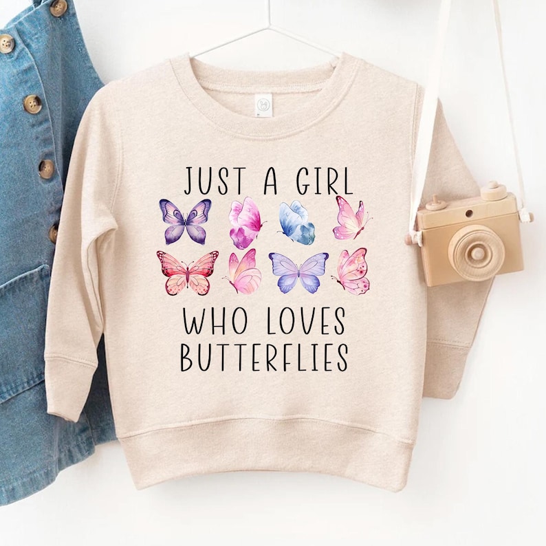 May include: A beige sweatshirt with the text "JUST A GIRL WHO LOVES BUTTERFLIES" in black. The design includes nine colorful butterfly illustrations in shades of purple, pink, and blue. The sweatshirt is hanging on a white hanger.