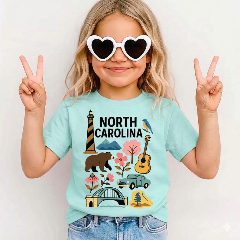 May include: A light blue t-shirt featuring a colorful graphic of North Carolina landmarks, including a lighthouse, mountains, a bear, a guitar, and a vintage car. The text "NORTH CAROLINA" is printed in black. The shirt is worn by a person wearing heart-shaped sunglasses.