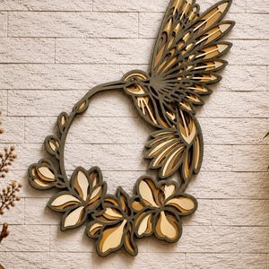 May include: A multi-layered wooden wall art piece featuring a hummingbird in flight integrated with a circular floral wreath. The design uses stacked layers of dark brown, tan, and cream wood to create depth, showcasing intricate cutouts of wings and petals.