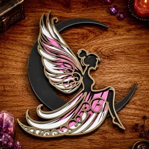Könnte beinhalten: Ein mehrschichtiges Wandkunstobjekt, das eine schwarze Silhouette einer geflügelten Figur zeigt, die auf einer Mondsichel sitzt. Das Design enthält weiße und rosa Akzente mit goldfarbenen Kanten, was durch filigrane Ausschnitte einen Effekt erzeugt.