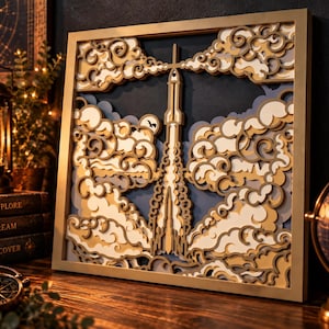 Op de afbeelding: Een vierkant, meerlaags houten wandkunstwerk met een centrale raket die omhoog stijgt door gestileerde, wervelende wolken. Het ontwerp gebruikt tinten goud, crème en donkerblauw, omlijst door een eenvoudige goudkleurige rand.