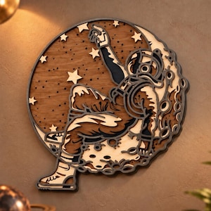 May include: A circular multi-layered wooden wall art piece featuring a detailed cutout of an astronaut sitting on a crescent moon. The design includes dark and light wood tones, depicting the astronaut reaching toward stars in a dark sky.