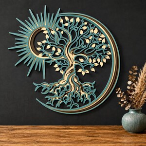 May include: A layered wooden wall art piece featuring a tree of life design integrated with a sun and crescent moon motif. The artwork uses teal, cream, and dark brown tones, creating a dimensional effect with intricate cutouts of branches and roots.