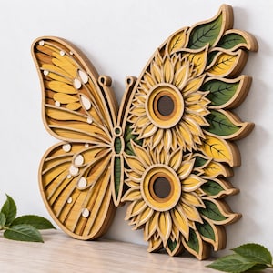 Puede incluir: Una pieza de arte mural de madera multicapa con forma de mariposa. El ala izquierda presenta patrones amarillos y blancos, mientras que la derecha se compone de dos girasoles con centros de color marrón oscuro y hojas verdes.